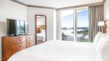 A hotel room with a large bed, a television on a wooden dresser, a balcony with a view of the city, and a sliding glass door.