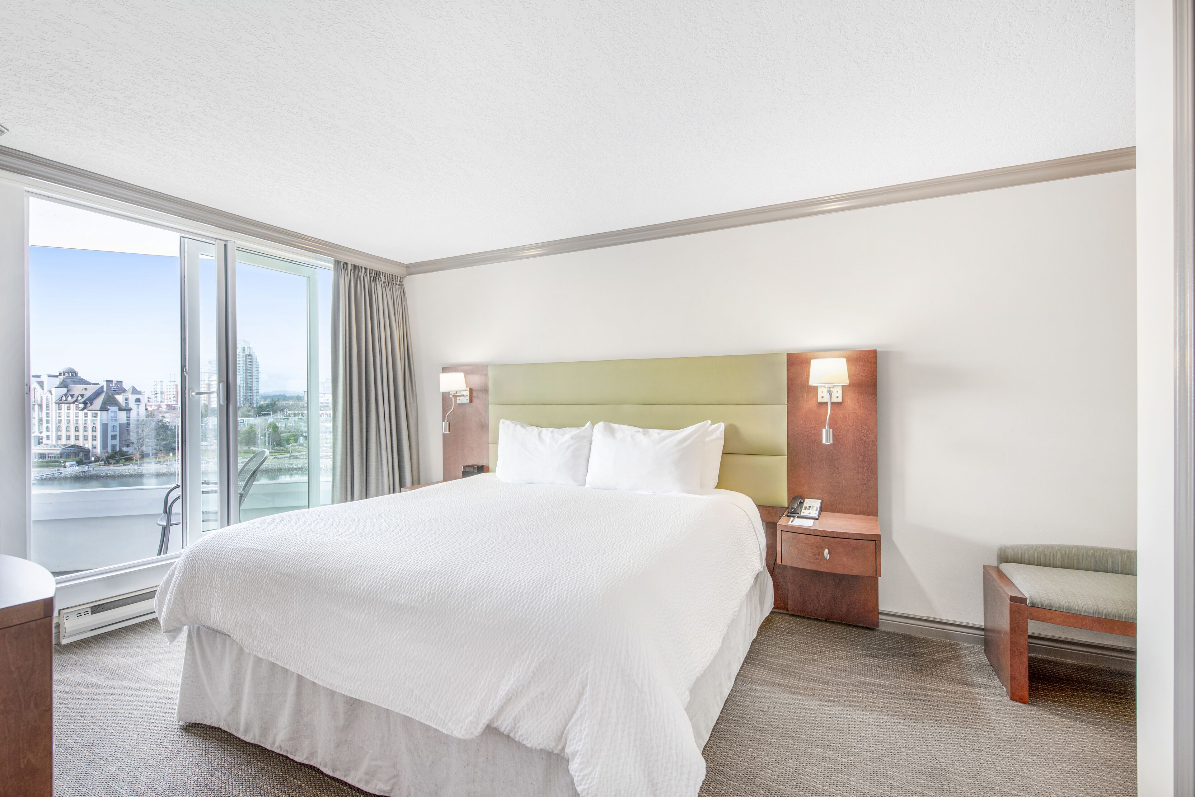 A hotel room with a large bed, a nightstand, a chair, and a view of the city through a large window.