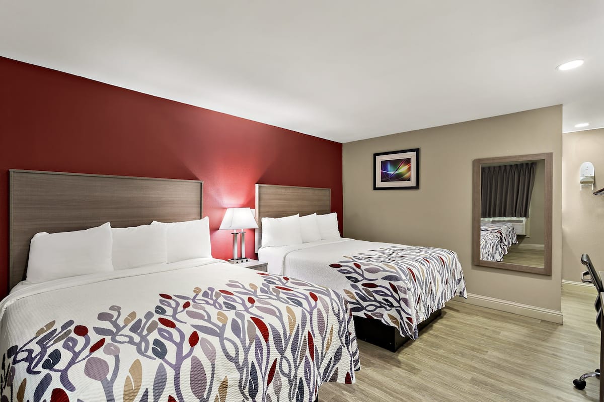 Quarto de hotel com duas camas, uma parede de destaque vermelha, um espelho e um abajur.