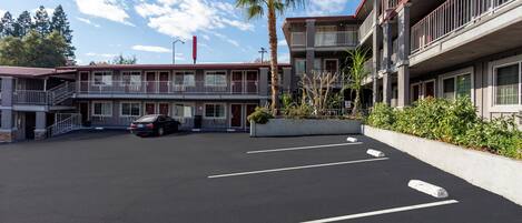 Un motel de varios pisos con estacionamiento, una palmera y cielo despejado.