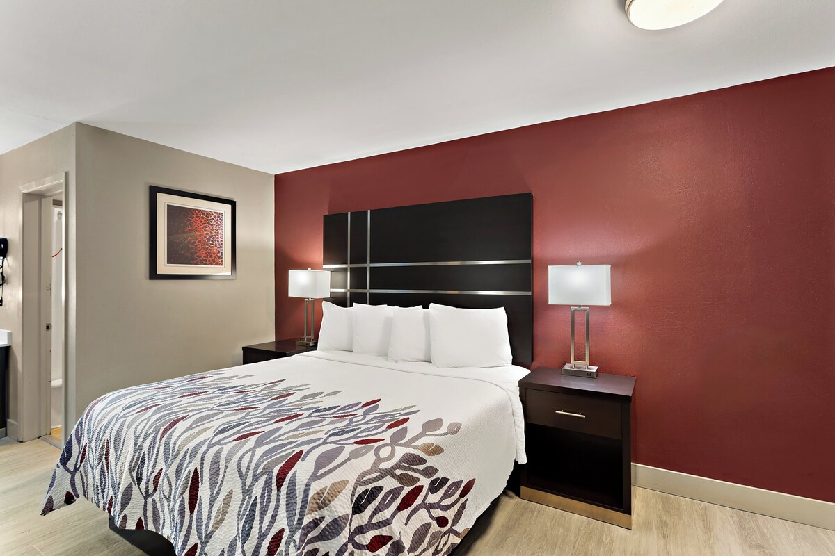 A hotel room with a large bed, a headboard, two bedside tables with lamps, a framed artwork on the wall, and a door to the left.