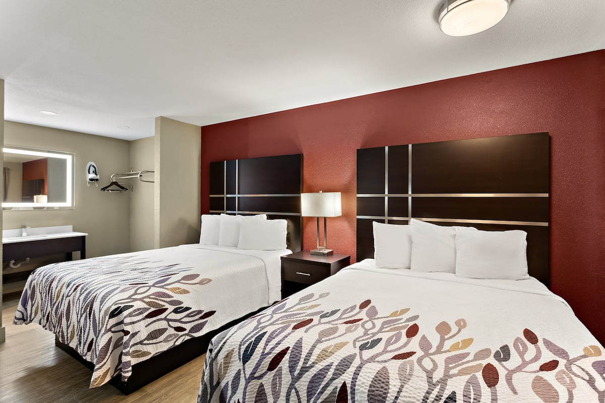 Quarto de hotel com duas camas, um espelho grande, cômoda e uma parede com destaque vermelha.
