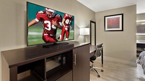 Une chambre d’hôtel dotée d’un grand téléviseur à écran plat fix é sur un meuble en bois, d’un bureau avec une chaise et d’une œuvre d’art abstraite encadrée accrochée au mur.