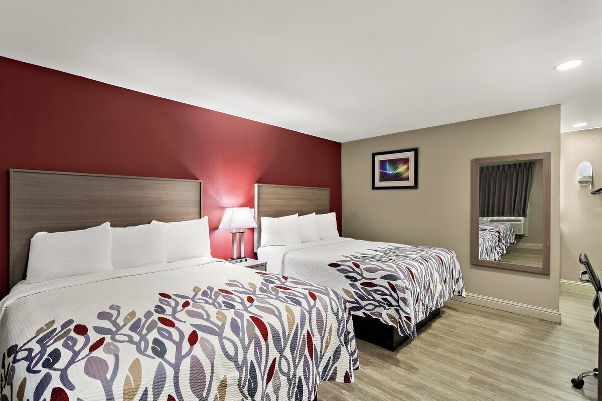 A hotel room with two beds, a red accent wall, a mirror, and a lamp.