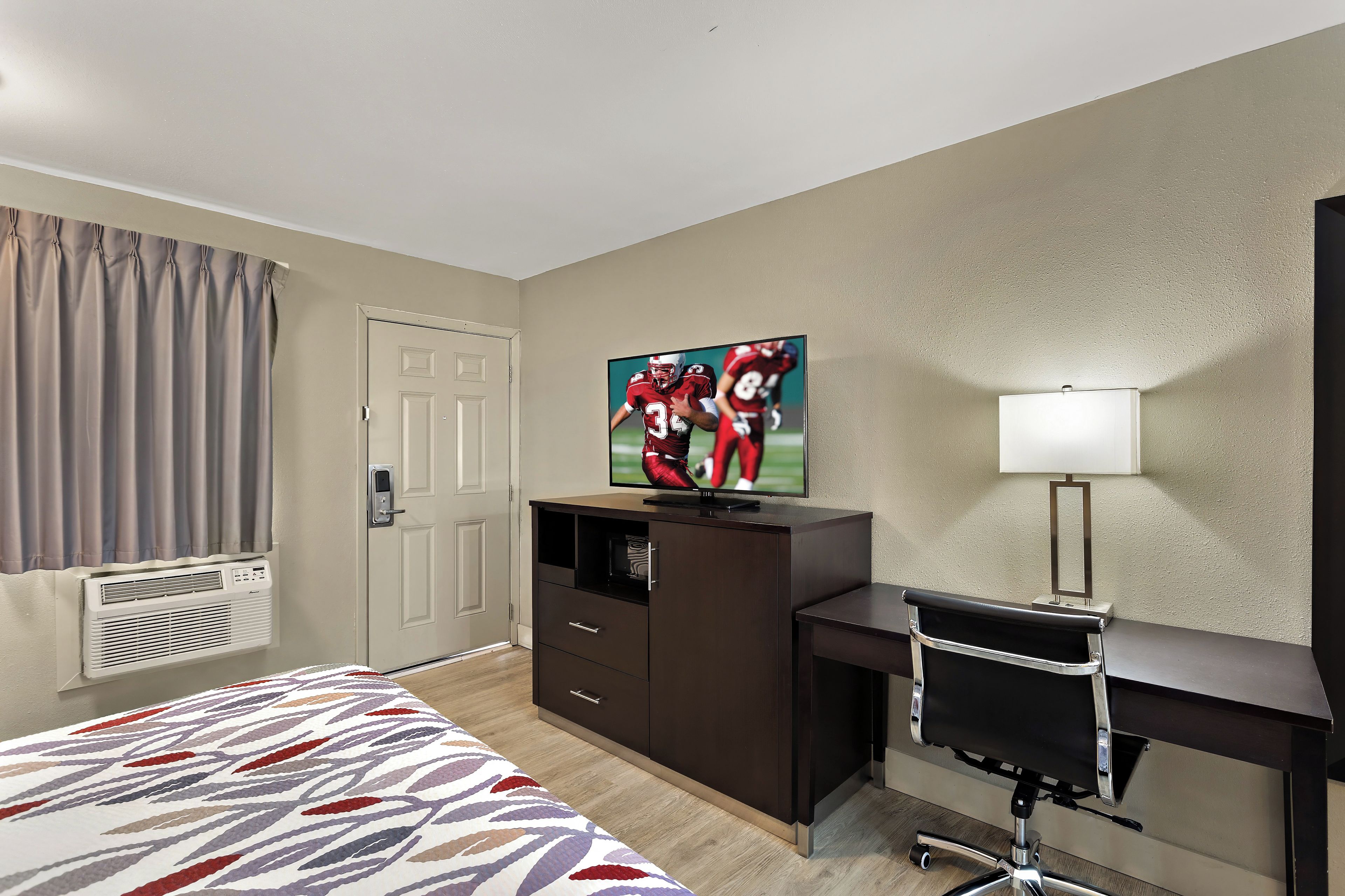A hotel room with a bed, a desk, a TV, a lamp, and a door.