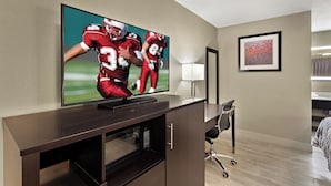 A hotel room with a flat-screen TV mounted on a wooden cabinet, a desk with a chair, a framed abstract painting on the wall, and a window with curtains. - Red Roof Inn Redding (Redding)
