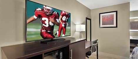 Habitación de hotel con una televisión de pantalla plana montada en un mueble de madera, un escritorio con una silla, un cuadro abstracto enmarcado en la pared y una ventana con cortinas.