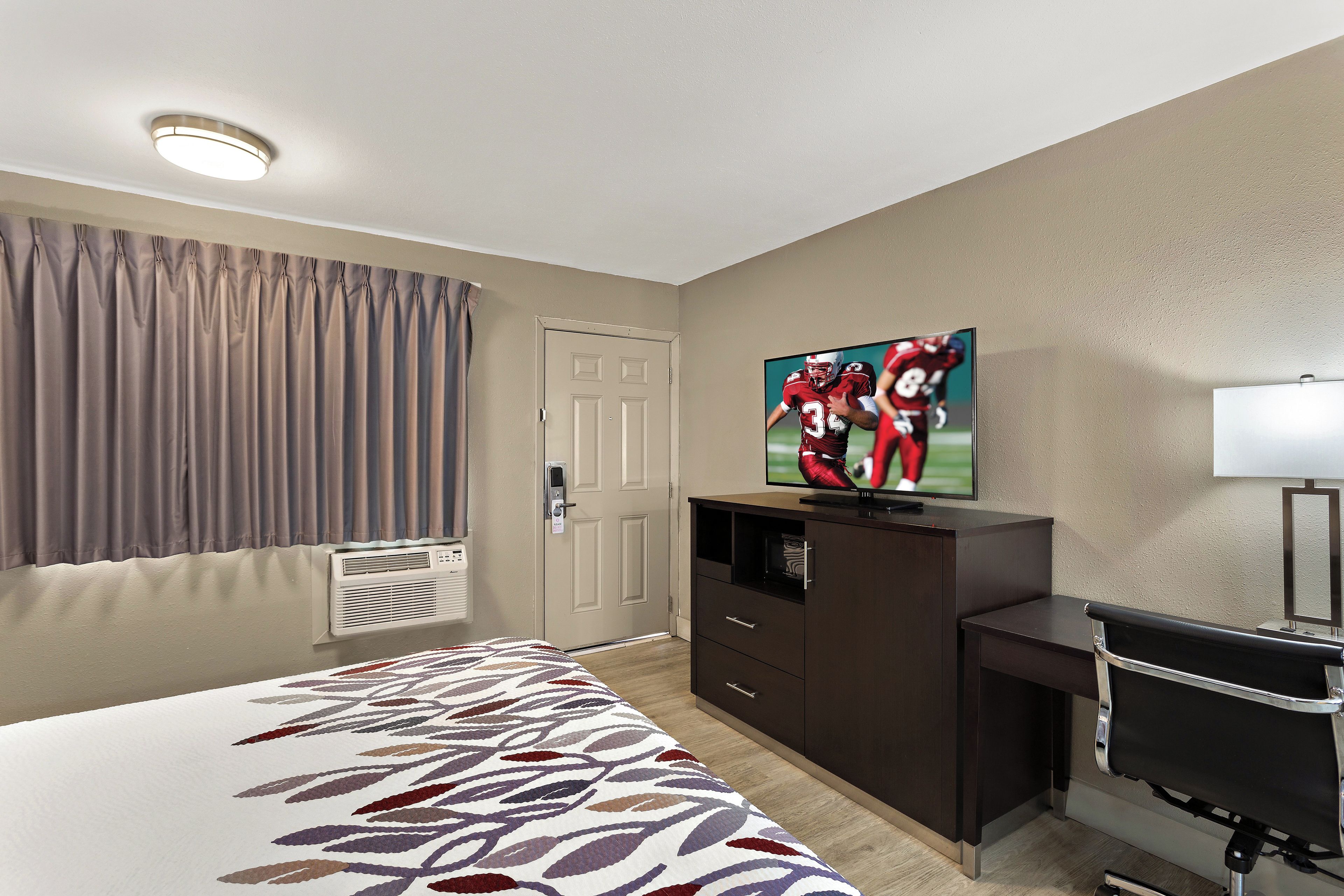 A hotel room with a bed, a television mounted on a dresser, a desk, and a window with curtains.