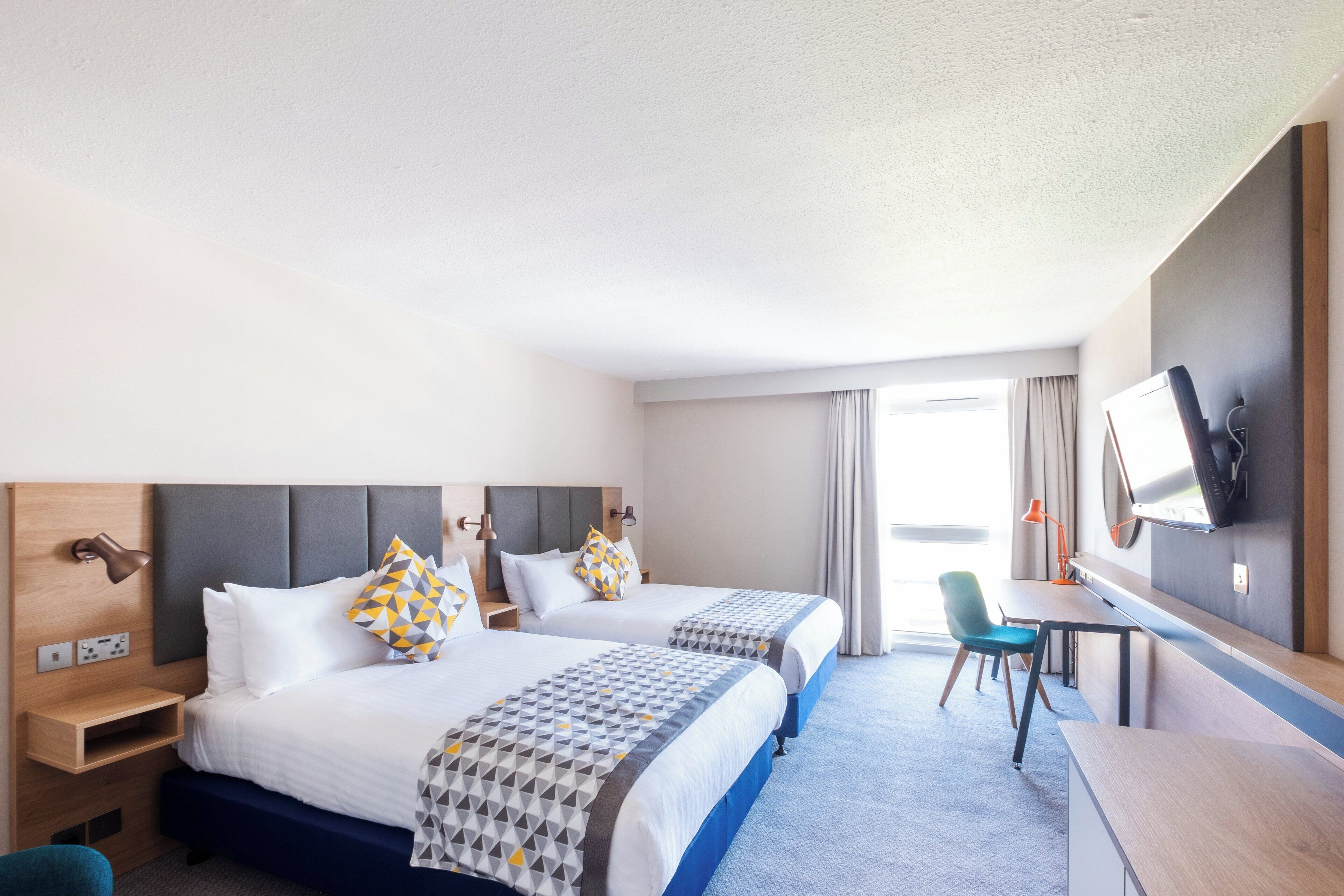 A hotel room with two beds, a desk, a chair, a TV, and a window with curtains.