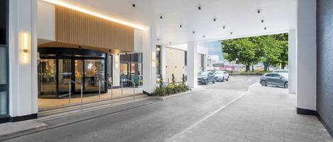 Una entrada de hotel moderna con fachada de vidrio, un área de estar al aire libre techada y un camino pavimentado que conduce a la entrada.