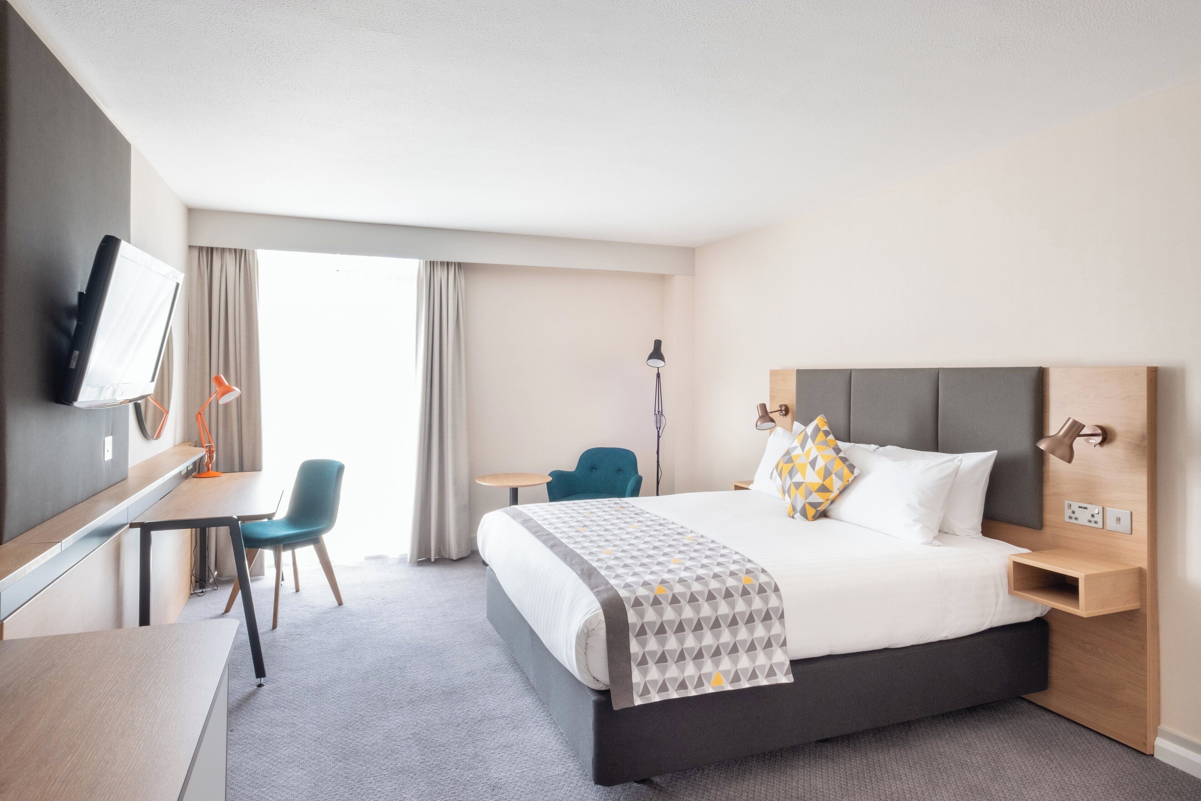 A hotel room with a bed, a desk, a chair, a TV, and a window with curtains.