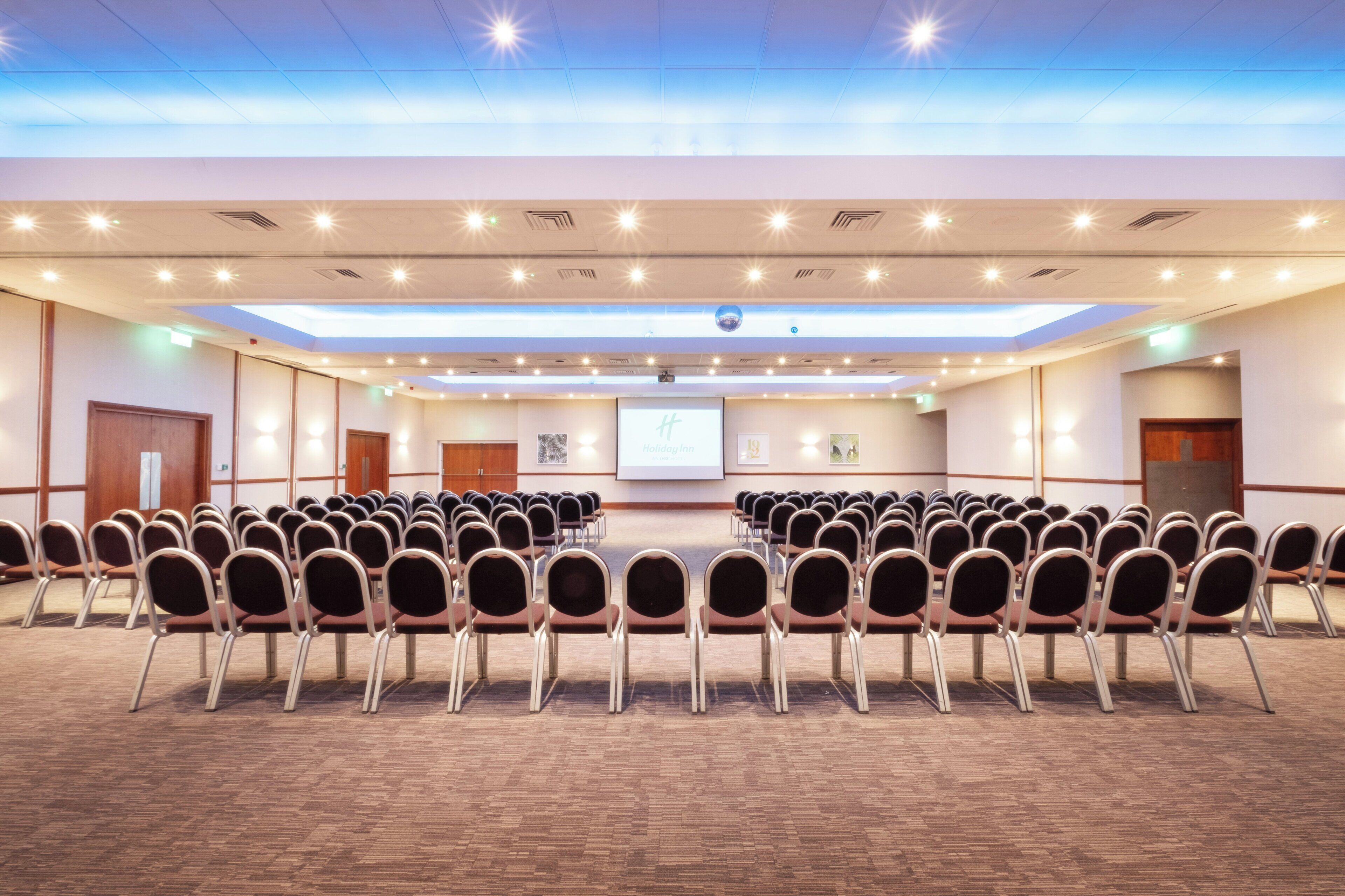 A conference room with rows of chairs, a projector screen, and a presentation on the wall.