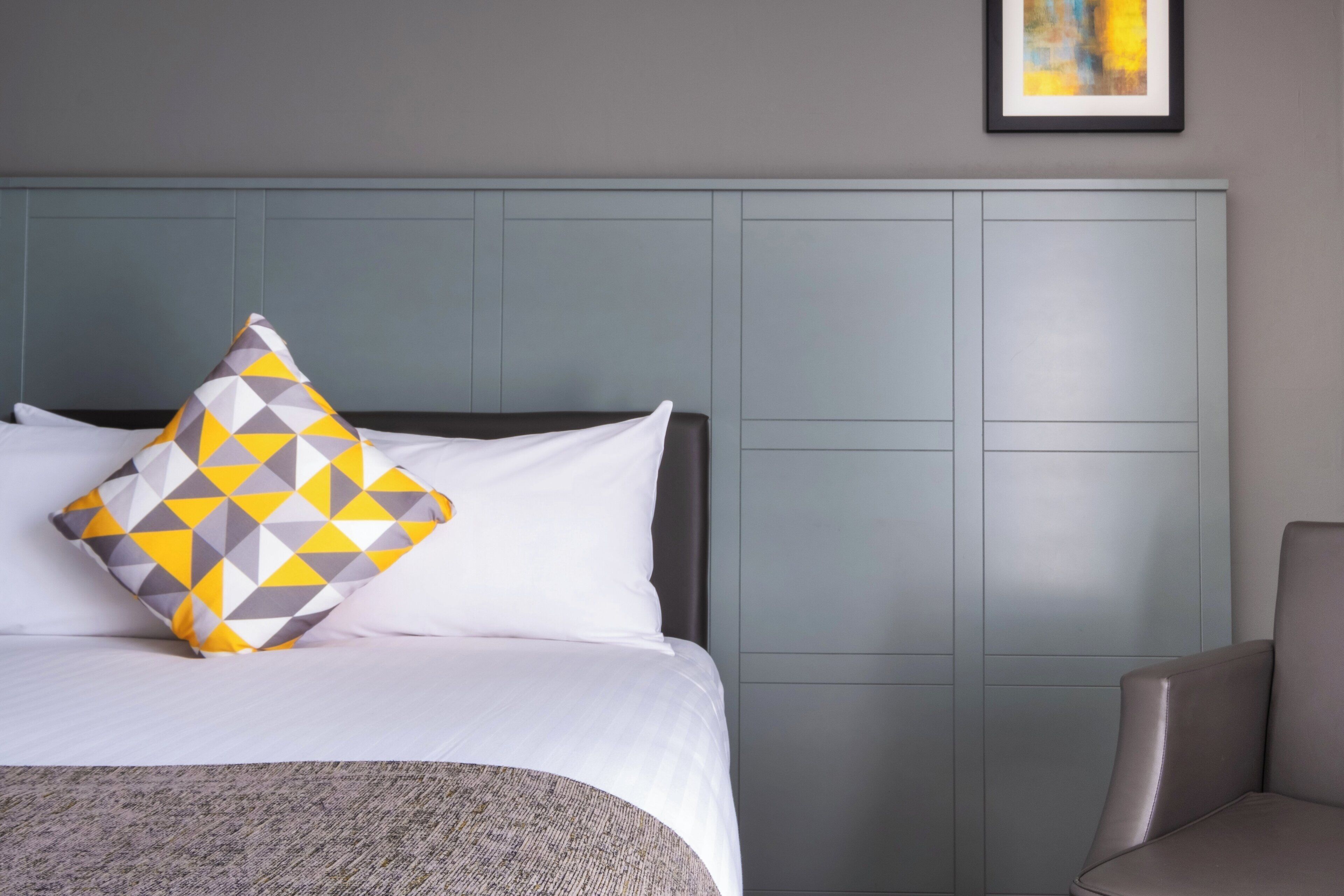 A hotel room with a bed, a grey headboard, a grey armchair, and a framed picture on the wall.