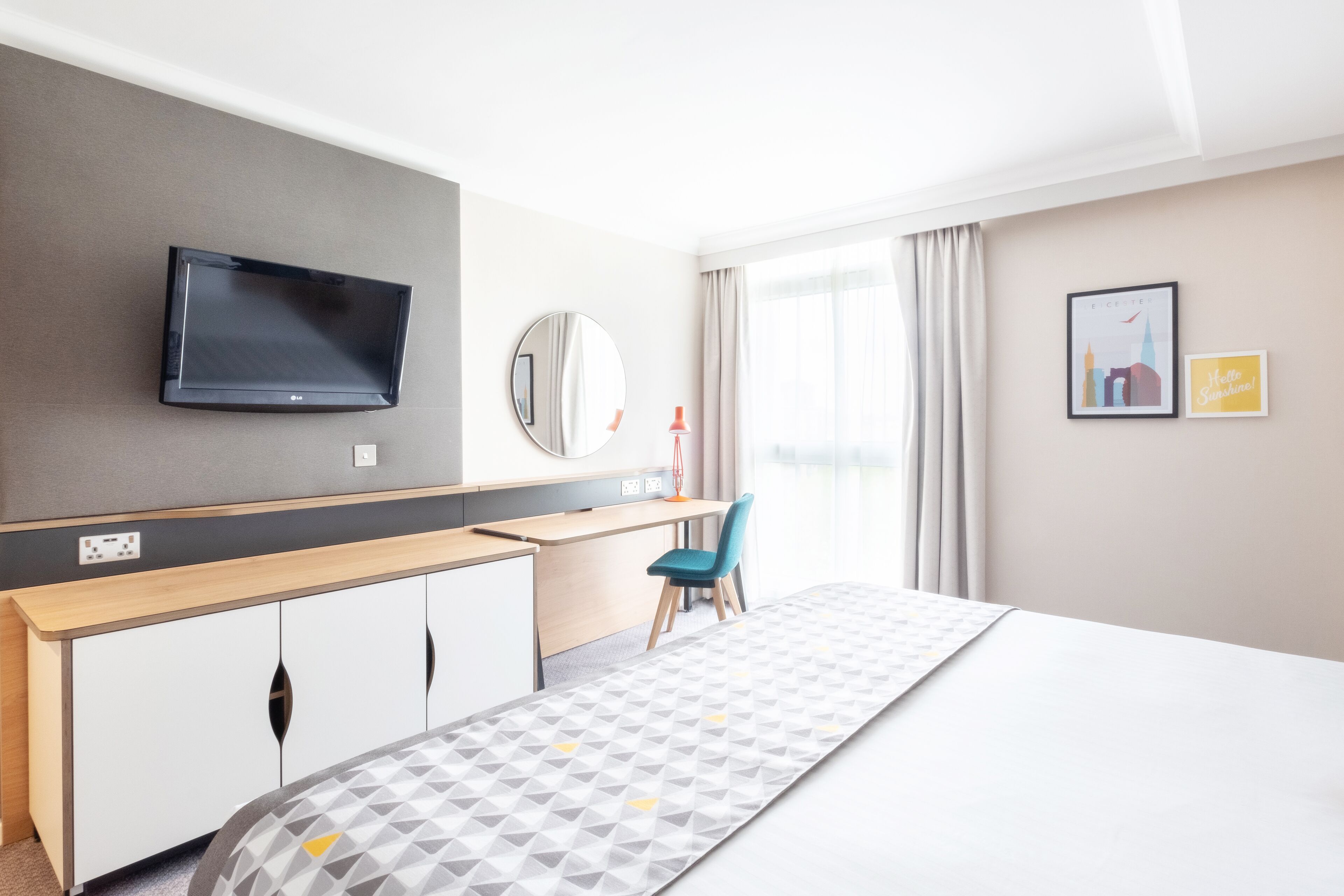 Una habitación de hotel moderna con una cama, un escritorio, una silla, un televisor, un espejo y una ventana con cortinas.