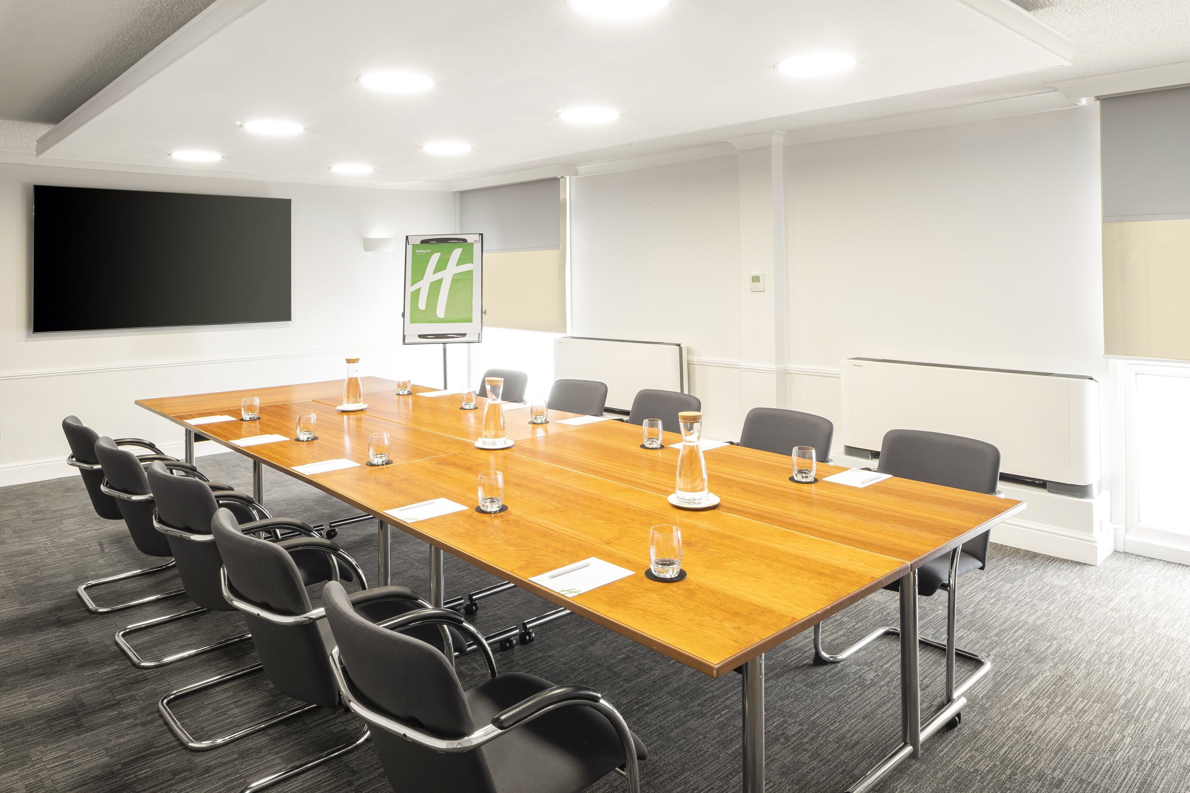 A conference room with a long table, black chairs, a projector screen, and a sign with a green logo.
