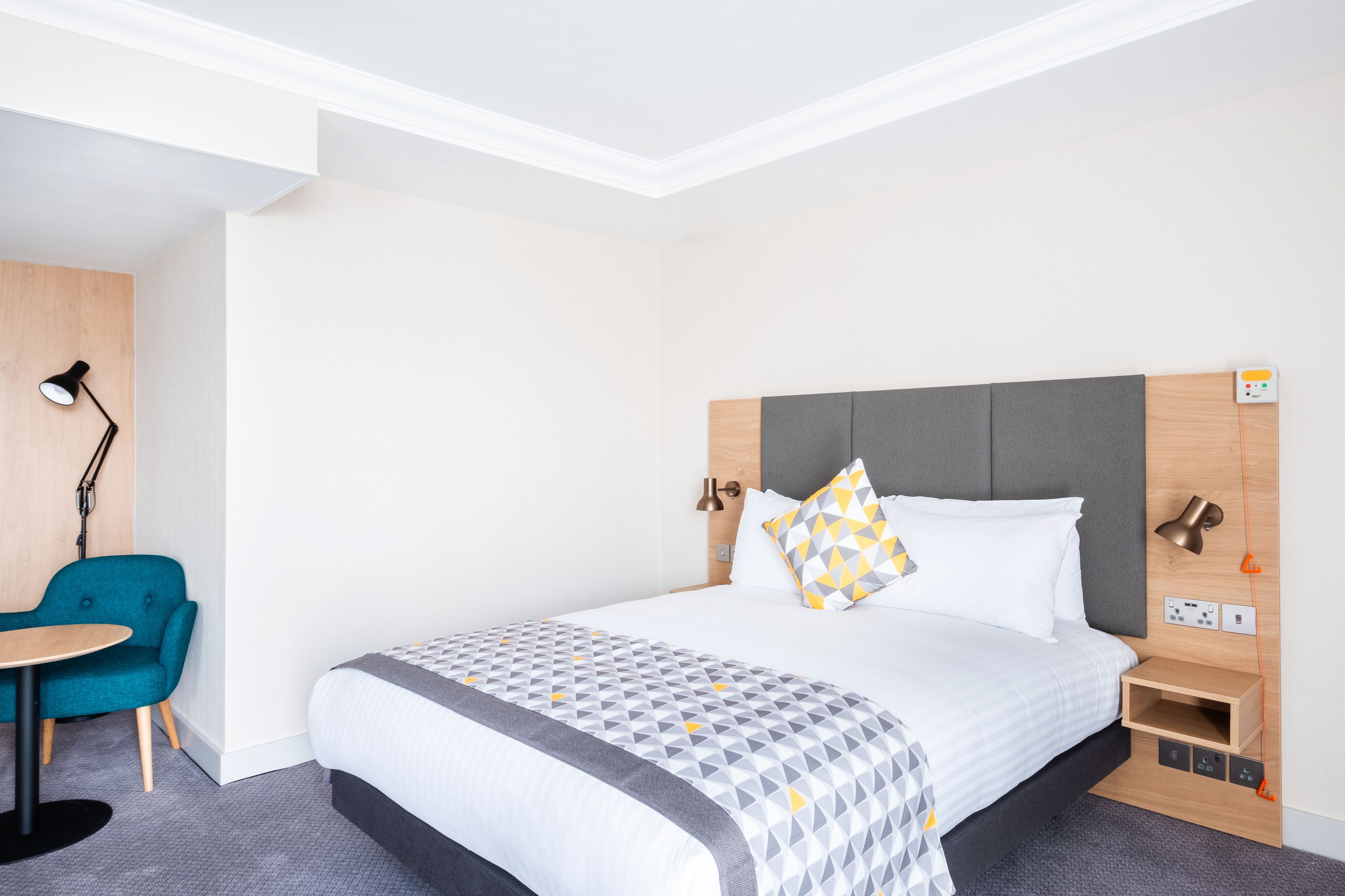 A hotel room with a bed, bedside table, lamp, chair, and a small table.