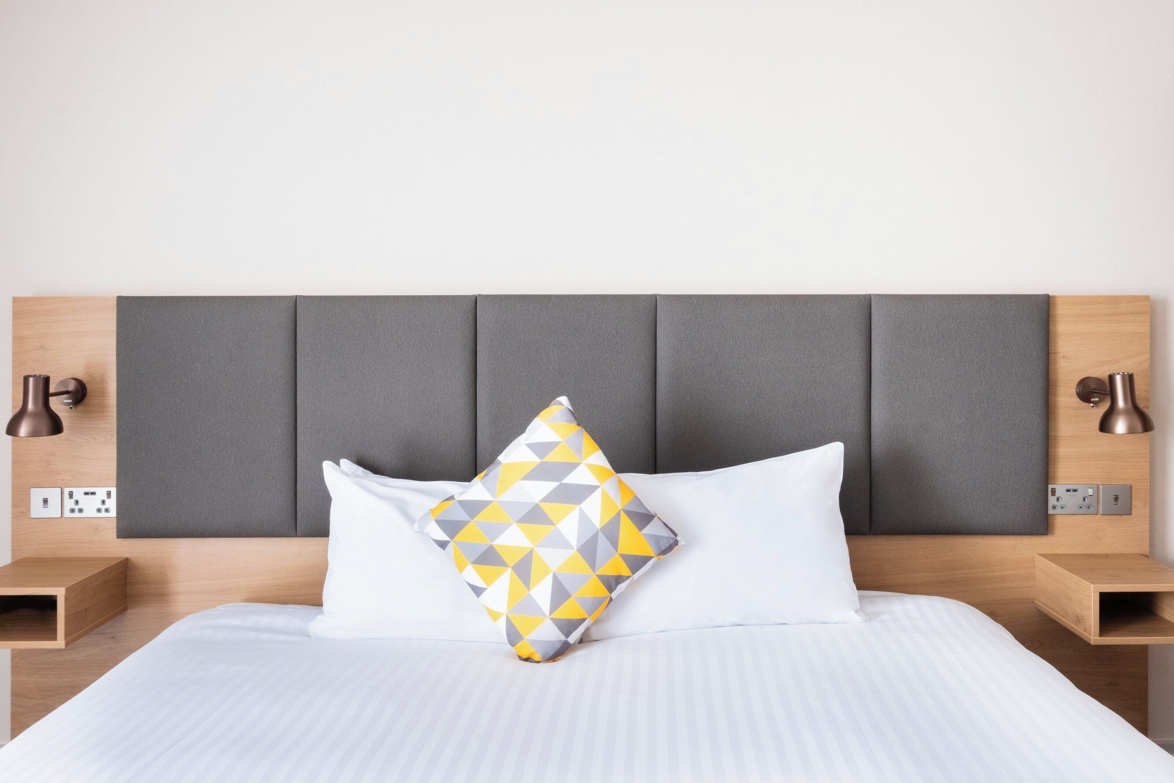 A neatly made bed with a grey headboard, two bedside lamps, and a power outlet on the wall.