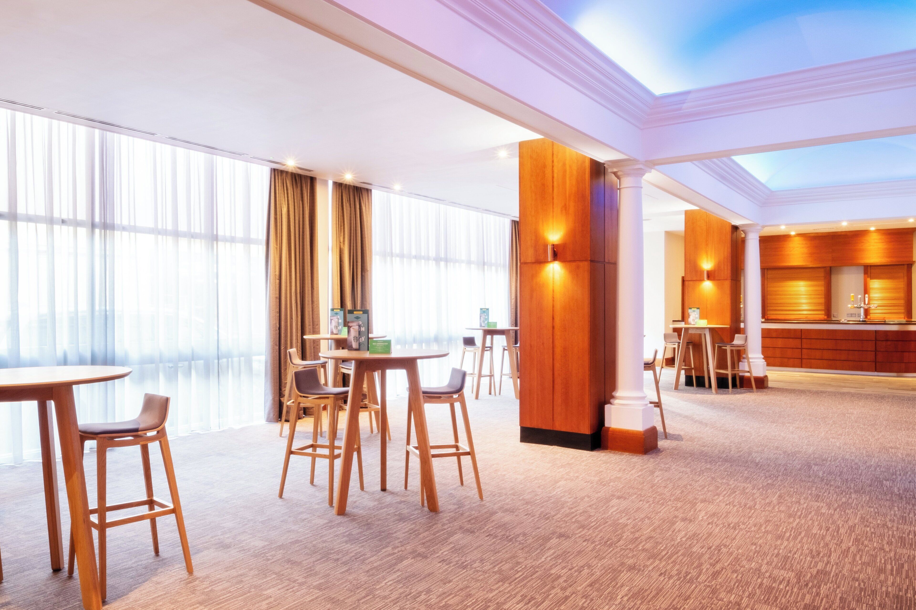 A hotel lobby with high ceilings, large windows, and a central seating area with tables and chairs.