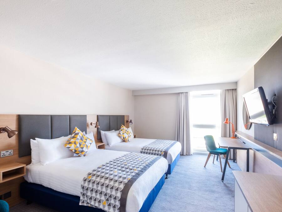Una habitación de hotel con dos camas, un escritorio, una silla, un televisor y una ventana con cortinas.