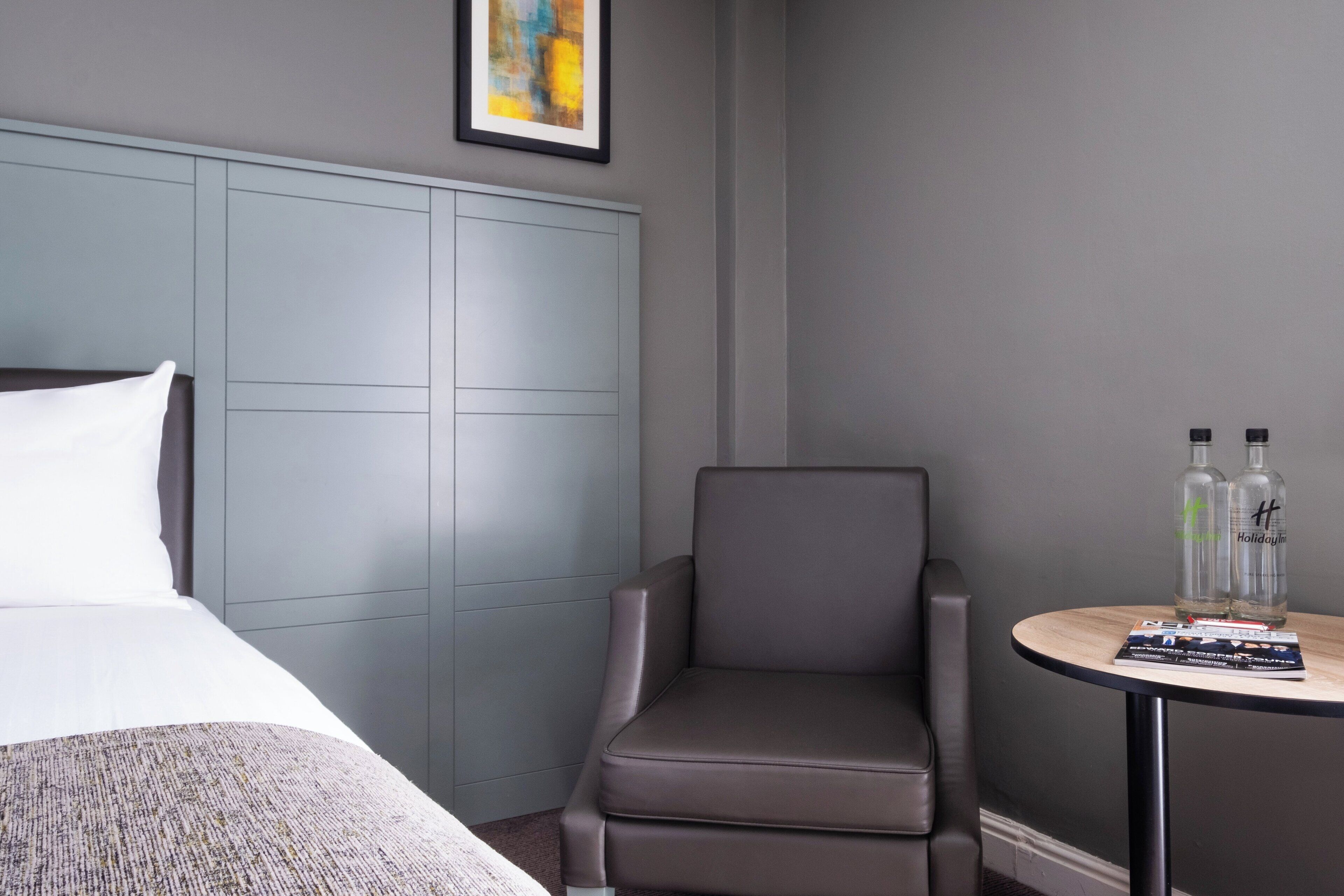 A hotel room with a bed, a grey upholstered chair, a small round table with two bottles, and a framed abstract painting on the wall.