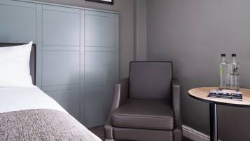 A hotel room with a bed, a grey upholstered chair, a small round table with two bottles, and a framed abstract painting on the wall.