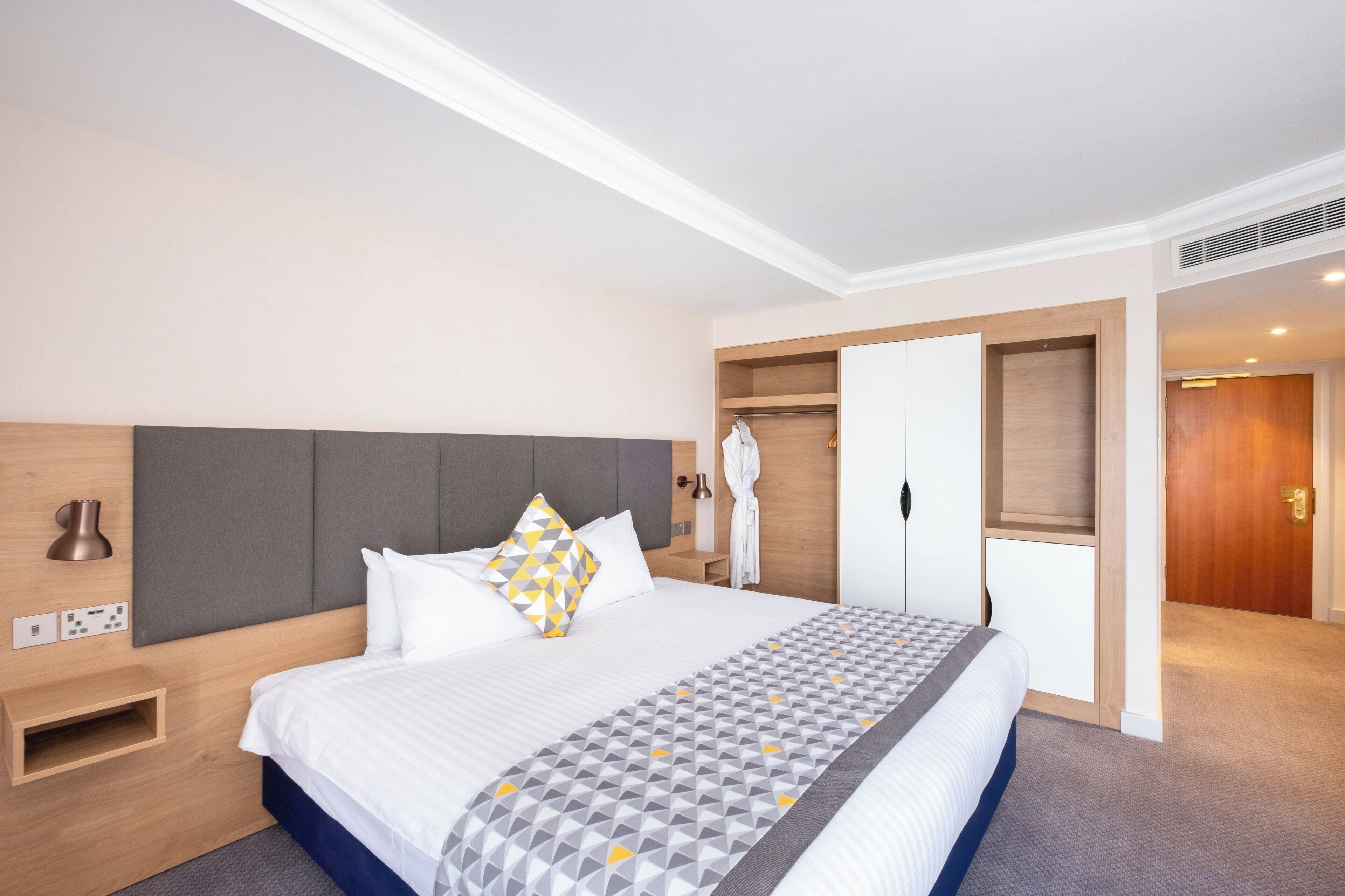 A hotel room with a bed, bedside table, wardrobe, and a bathroom visible through an open door.