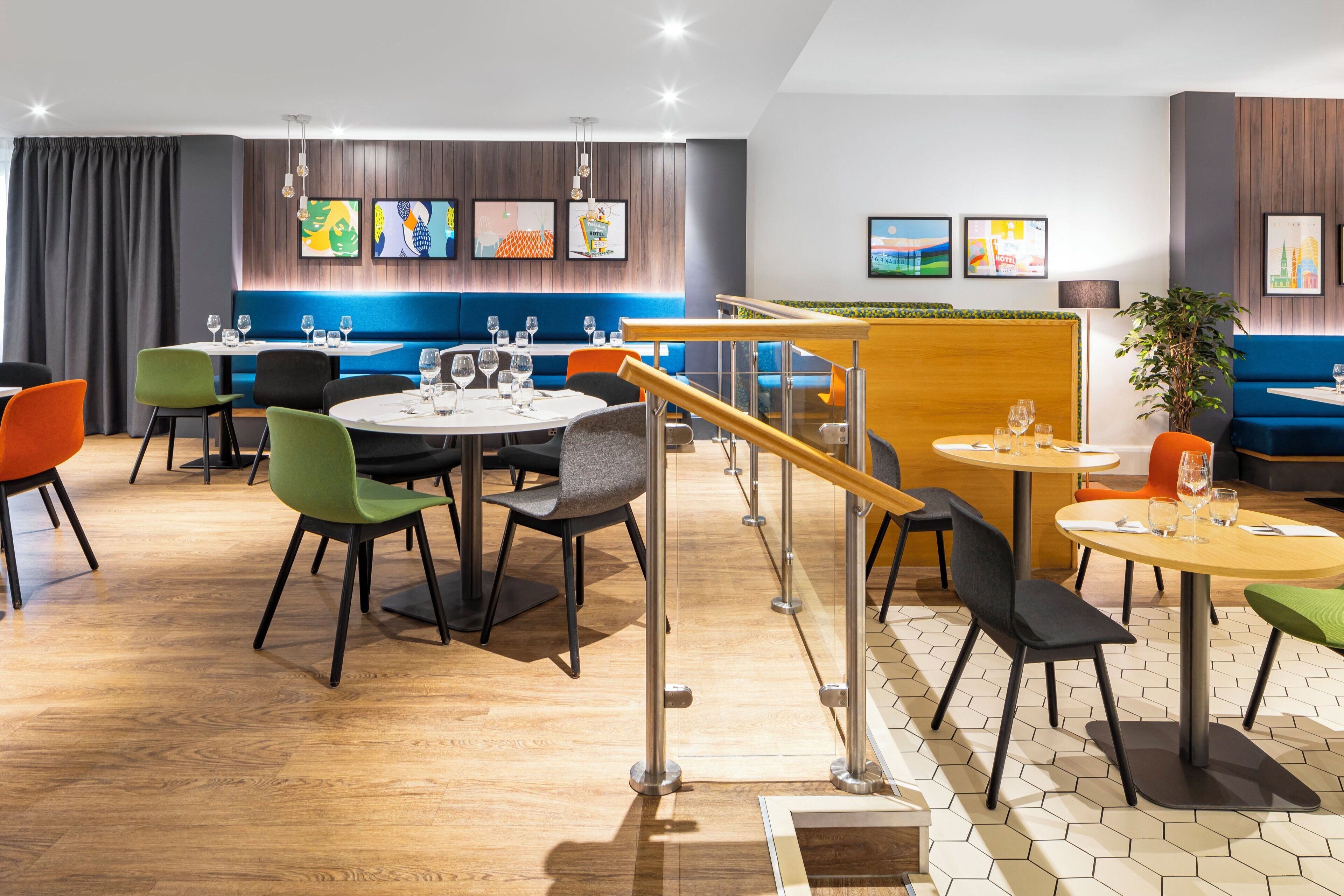A modern restaurant with round tables and colorful chairs, a wooden staircase, and artwork on the walls.