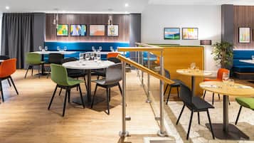 A modern restaurant with round tables and colorful chairs, a wooden staircase, and artwork on the walls.