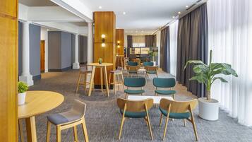 Um lobby de hotel com design moderno, apresentando uma combinação de mobiliário de madeira e estofado, uma área de estar central e uma grande planta em vaso.