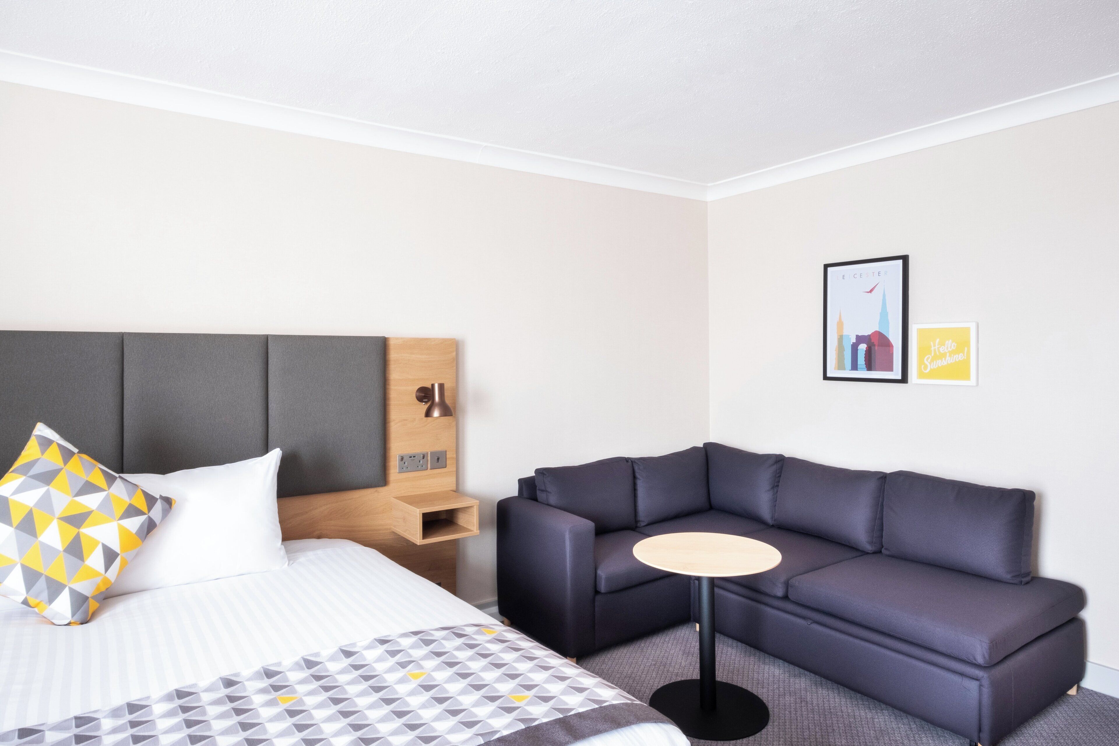A hotel room with a bed, a dark-colored sofa, a small round table, and a wall-mounted lamp.