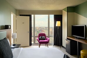 Quarto de hotel com cama, escrivaninha com televisão, cadeira e vista da cidade por grandes janelas.