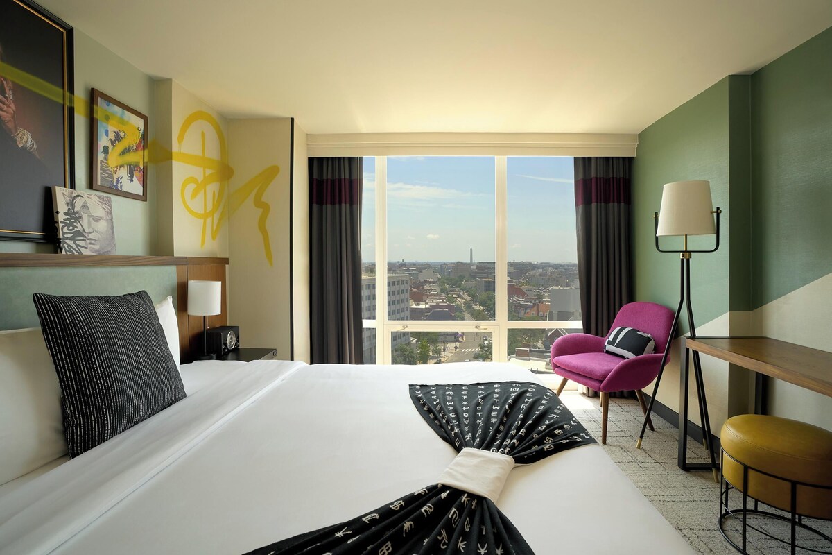 A hotel room with a large bed, a desk, a chair, a lamp, and a view of the city.
