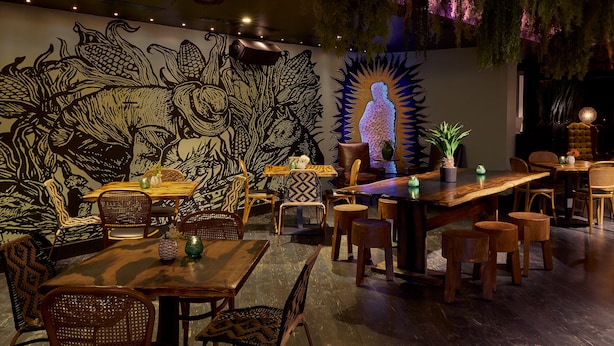 A restaurant interior with wooden tables and chairs, a mural on the wall, and hanging plants.