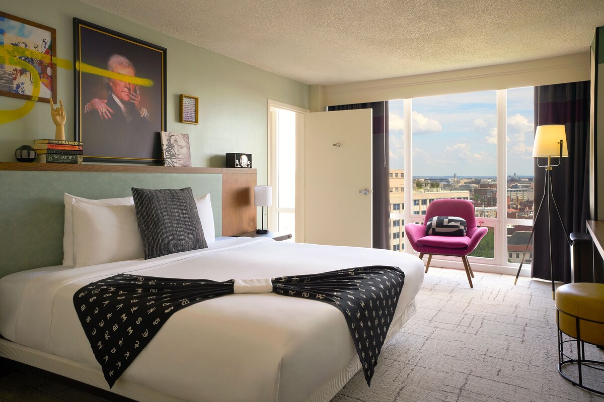 A hotel room with a large bed, a pink chair, a yellow stool, and a view of the city through the window.