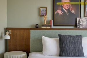 Un cuarto de hotel con cabecera de madera, una mesita de noche redonda, un almohadón blanco, un almohadón con estampado blanco y negro, una cita enmarcada en la pared y un retrato enmarcado.
