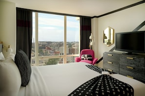Una habitación de hotel con un ventanal grande, una cama, un televisor, un aparador y una silla.