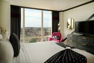 A hotel room with a large window, a bed, a television, a dresser, and a chair.