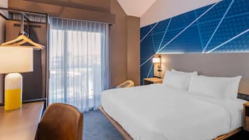 Une chambre d’hôtel comprenant un lit, un bureau avec une lampe, une chaise, une armoire et une grande fenêtre avec des rideaux.