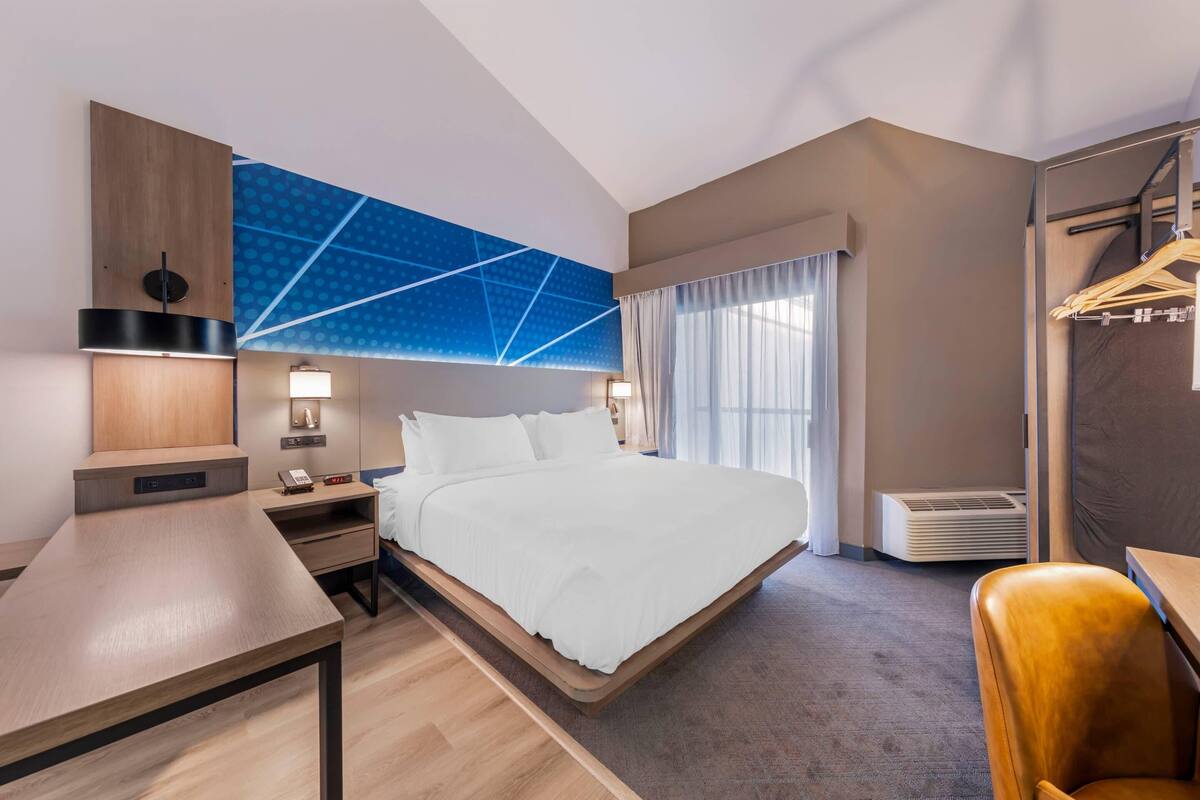 Moderni hotellihuone, jossa on sänky, yöpöydät, työpöytä, vaatekaappi ja suuri ikkuna, jossa on verhot.