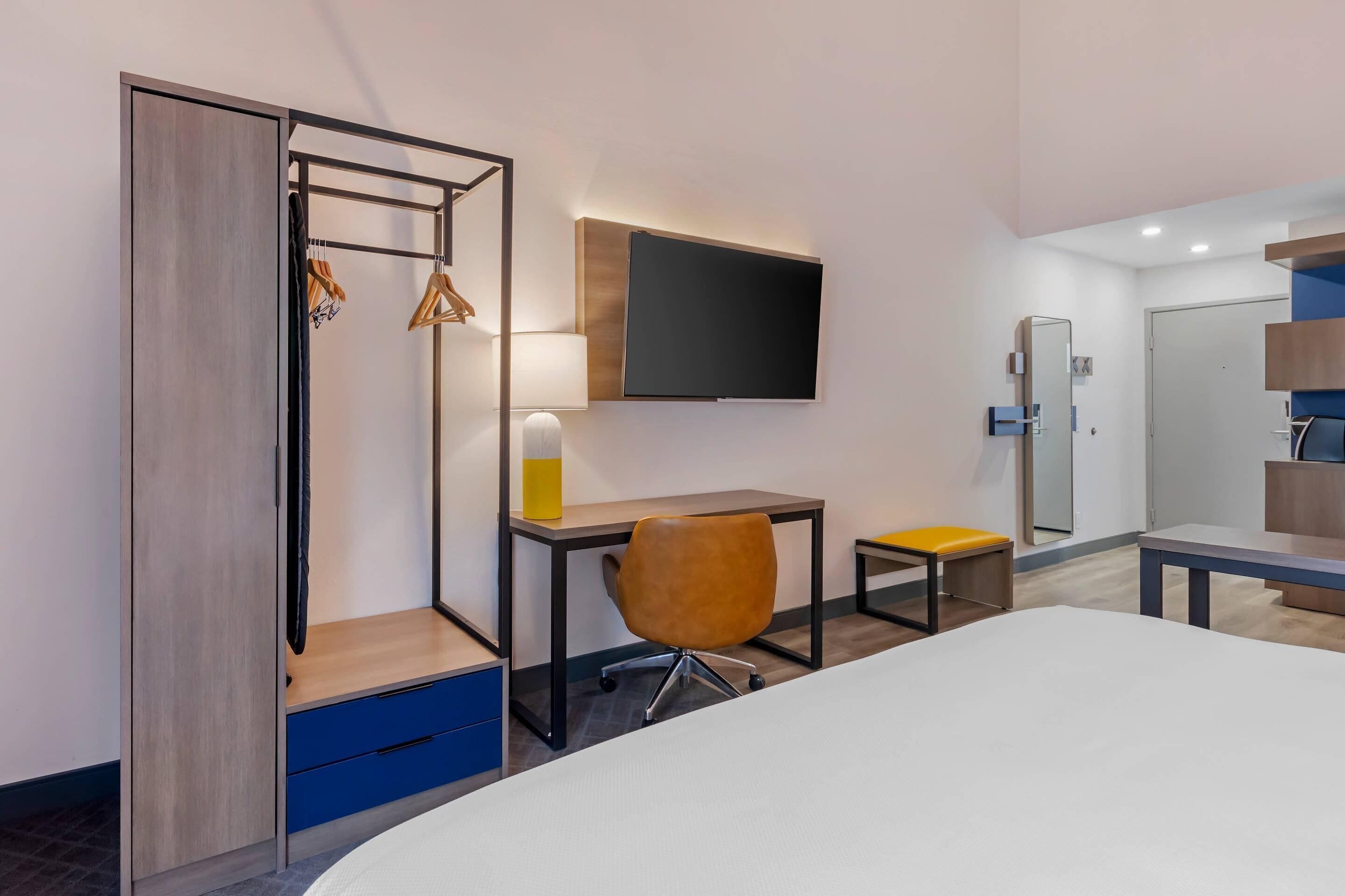A hotel room with a bed, a desk with a chair, a TV mounted on the wall, and a wardrobe with hanging clothes.