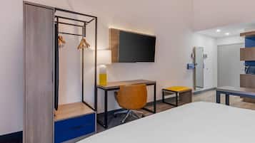 A hotel room with a bed, a desk with a chair, a TV mounted on the wall, and a wardrobe with hanging clothes.