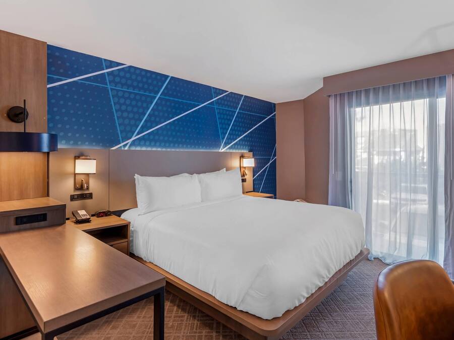 Une chambre d’hôtel comprenant un lit, des tables de chevet, un bureau, une horloge murale et une grande fenêtre avec des rideaux.