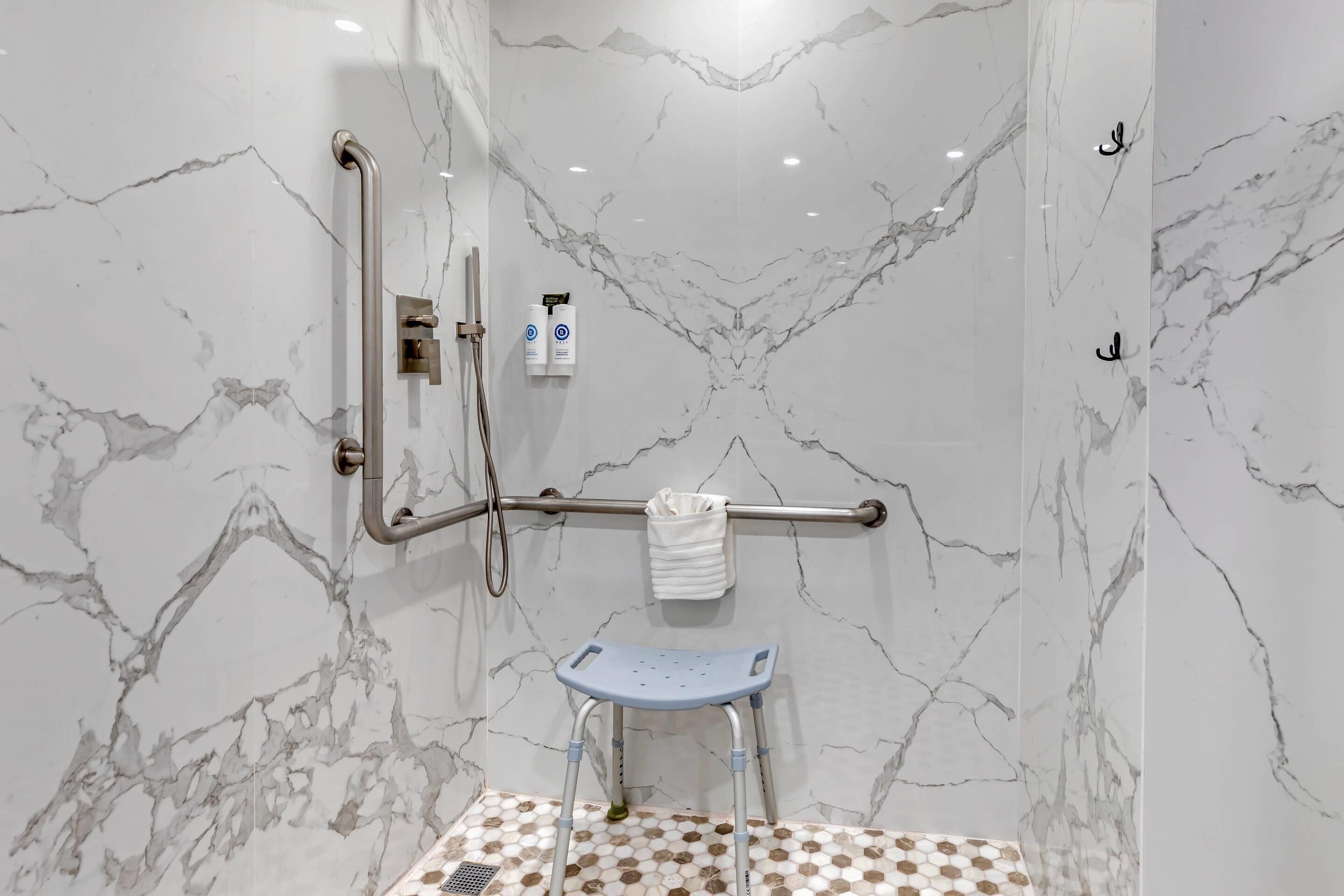Un baño con paredes de mármol, ducha con cabezal de ducha manual, barra de apoyo y un taburete.