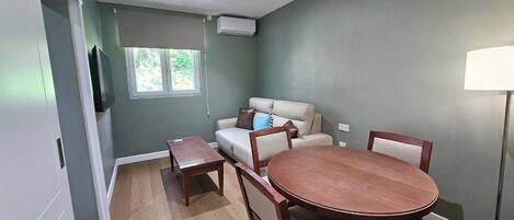 Uma área de refeições com uma mesa redonda de madeira, quatro cadeiras, um sofá e uma TV fixada na parede.