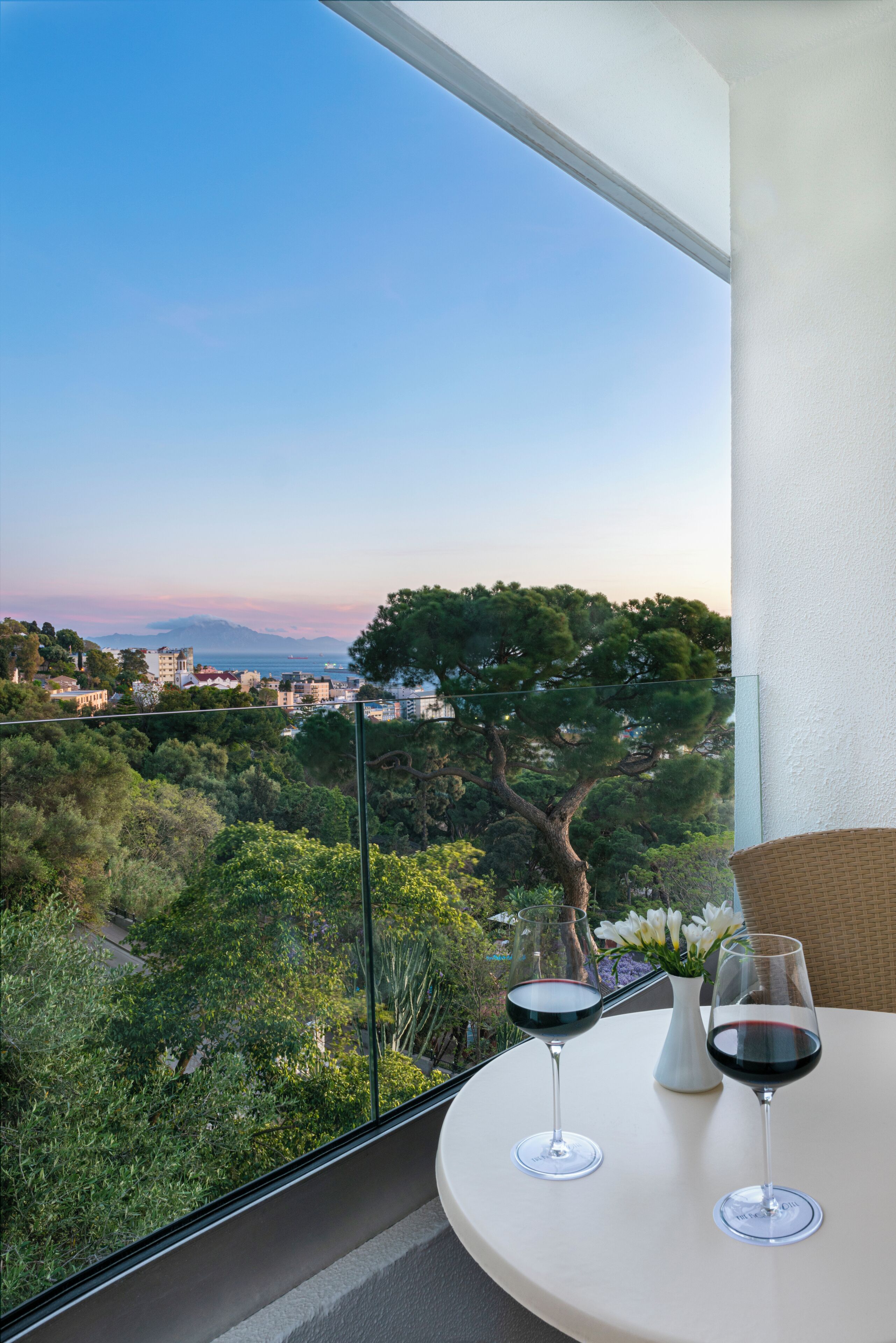 A balcony with two wine glasses, a wicker chair, and a view of a coastal town with trees and buildings.