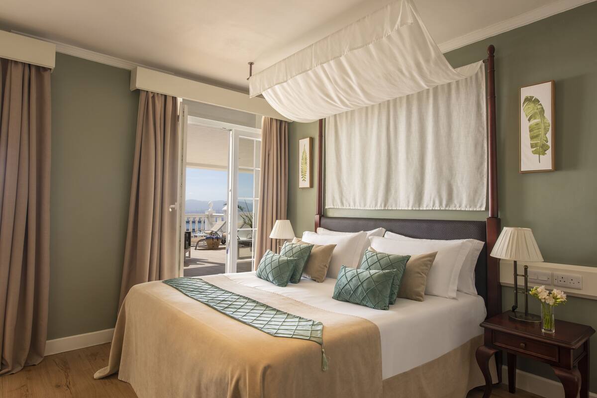 A hotel room with a large bed, a canopy bed, a bedside table with a lamp, and a view of the outdoors.