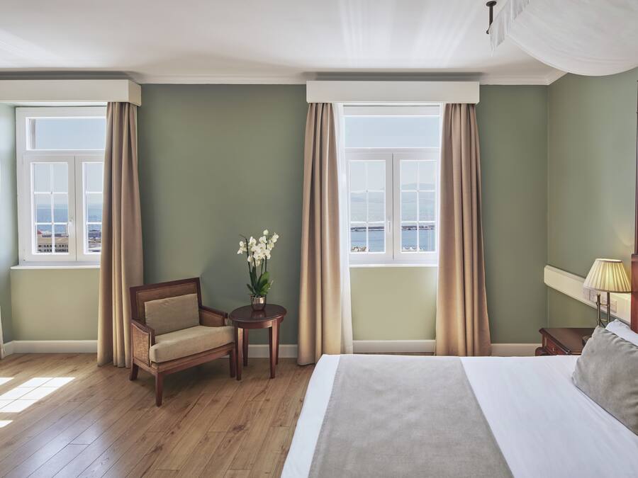 Une chambre d’hôtel avec un grand lit, une chaise, une petite table ornée d’un vase de fleurs et une vue sur la mer grâce à de grandes fenêtres.