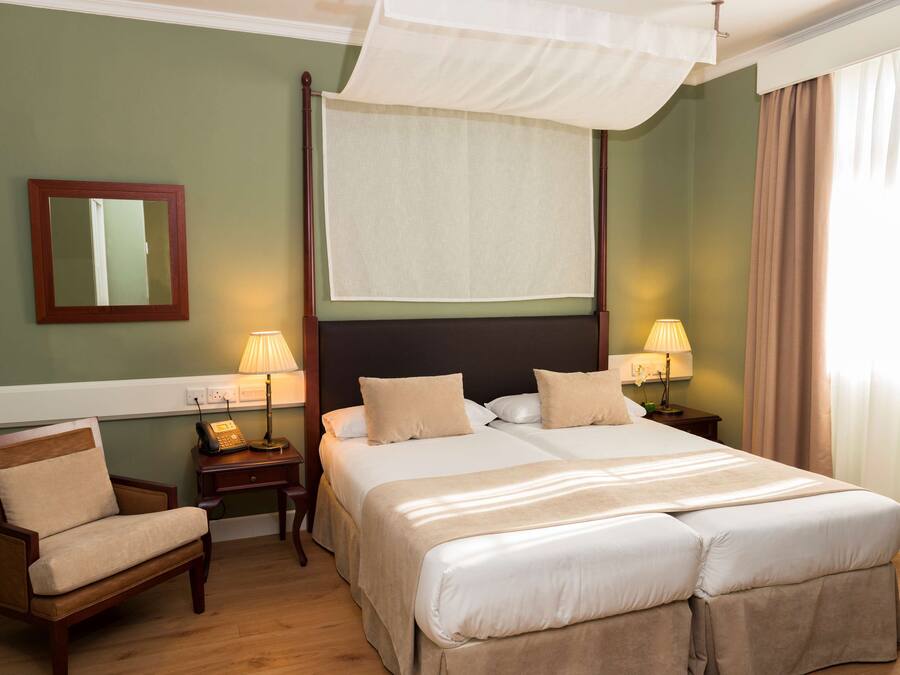 Habitación de hotel con una cama grande, dos mesitas de noche con lámparas, una silla, un espejo y un dosel.