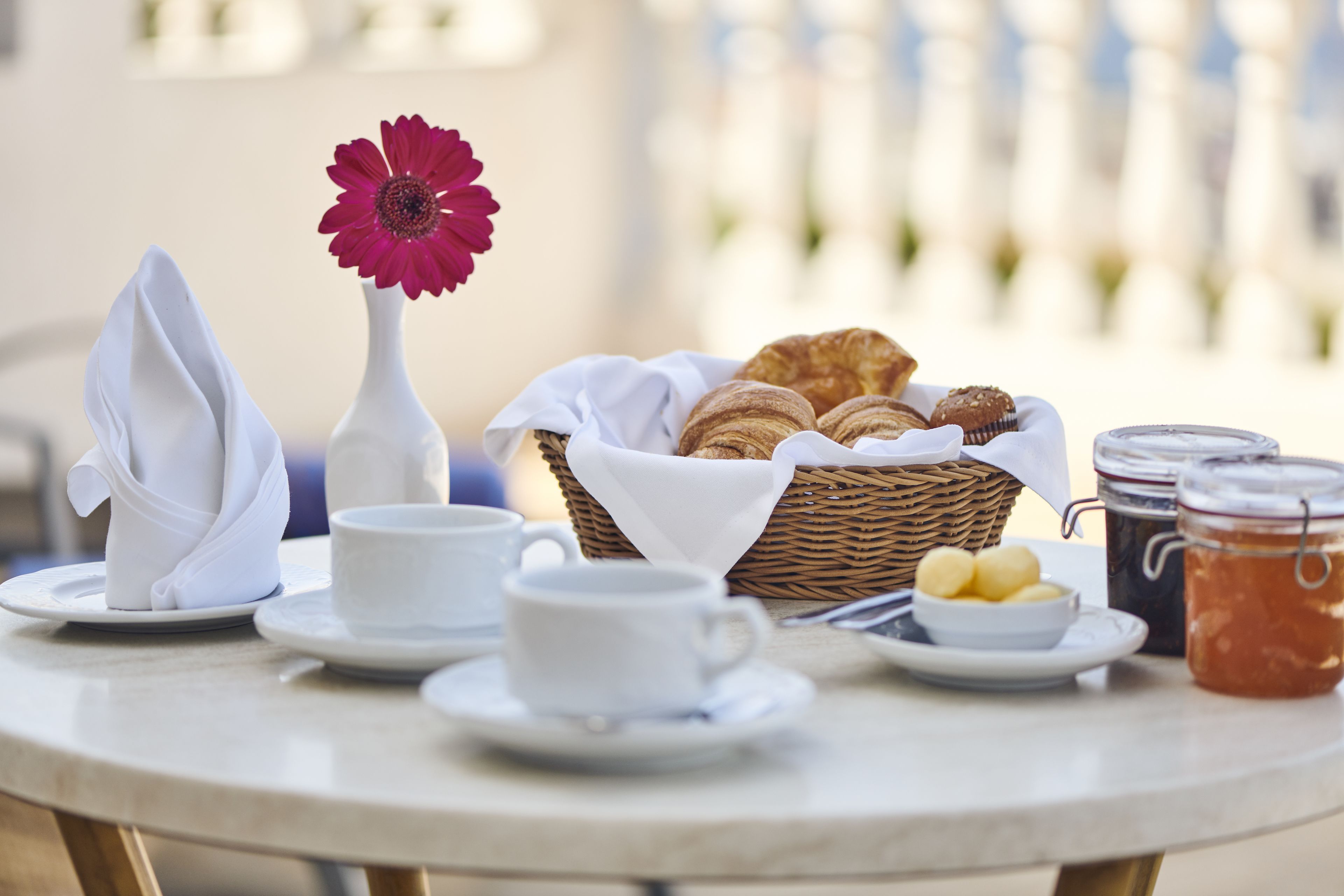 Ein Frühstückstisch mit einem Korb voller Croissants, einer Vase mit einer einzelnen Blume und zwei Kaffeetassen.