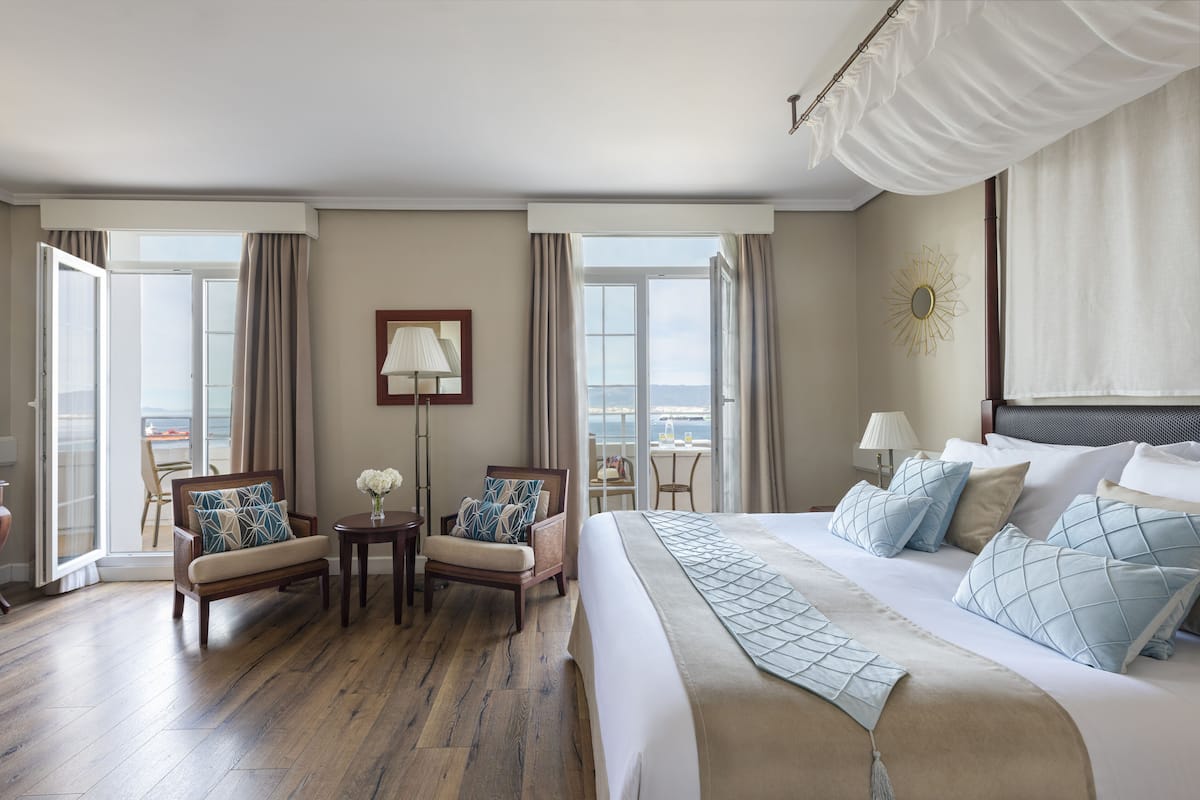 A hotel room with a large bed, two armchairs, a small table, and a view of the sea through large windows.